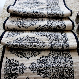 May include: A white table runner with a black damask pattern and black trim. The runner is folded in fourths and is lying on a white fluffy surface.