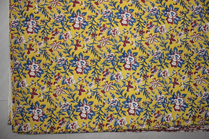 May include: A vibrant yellow fabric featuring a dense, repeating floral pattern. The design consists of small white flowers with dark red centers, interspersed with blue leaves and thin, winding stems that cover the entire surface of the textile.