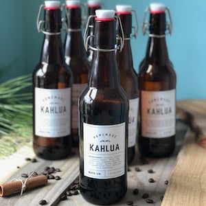 May include: Six brown glass bottles with white labels reading "Homemade Kahlua." The bottles have swing-top closures and are filled with a dark liquid. Coffee beans and a cinnamon stick are on a wooden surface.