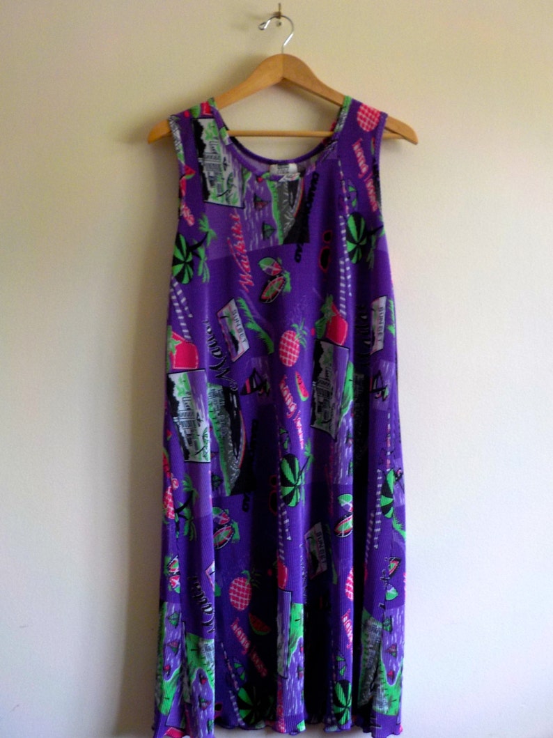 May include: A purple sleeveless maxi dress with a colorful print of tropical fruits, palm trees, and geometric shapes. The dress has a relaxed fit and a flowy silhouette.