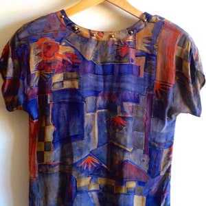 May include: A short-sleeved, abstract print blouse with a geometric pattern in shades of blue, brown, red, and gold. The neckline is adorned with a row of gold studs.