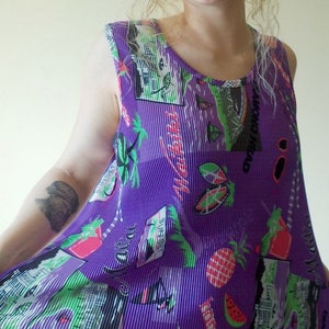 May include: Purple sleeveless top with a pleated design and a colorful print of palm trees, pineapples, watermelons, and surfboards. The print also includes the text "Miami Beach" and "South Street Seaport".