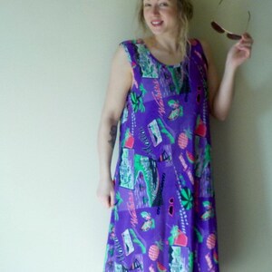 May include: A woman wearing a purple, sleeveless dress with a colorful tropical print. The dress has a loose fit and falls to her knees. She is wearing a pink headband and has sunglasses in her hand.