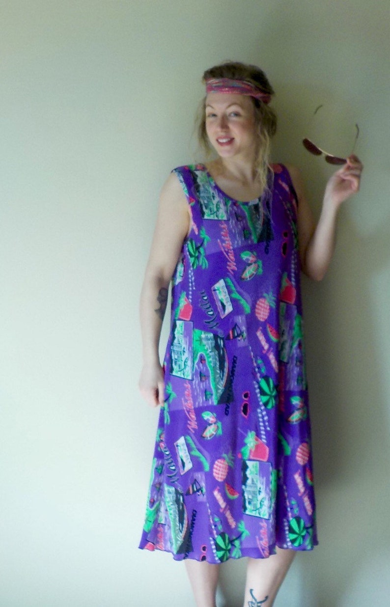 May include: A woman wearing a purple, sleeveless dress with a colorful tropical print. The dress has a loose fit and falls to her knees. She is wearing a pink headband and has sunglasses in her hand.