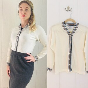 May include: A white cardigan sweater with a striped trim around the neckline and cuffs. The cardigan has a button closure and is made of a soft, lightweight fabric.