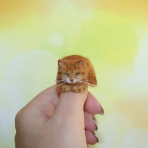 Op de afbeelding: Een klein, oranje en wit vilten kattenbeeldje, slaapt op een vinger.