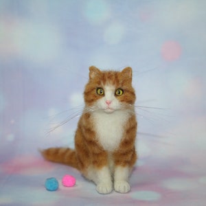 May include: A needle-felted orange and white tabby cat figurine with yellow eyes, a pink nose, and long thin whiskers. The cat is sitting upright with its tail extended to the side, accompanied by one small blue and one small pink felted ball.
