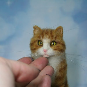 May include: A needle felted cat figurine featuring orange and white fur, pointed ears, and bright yellow eyes. The cat has a small pink nose and long, thin, clear whiskers extending from its muzzle, crafted with a textured, soft appearance.