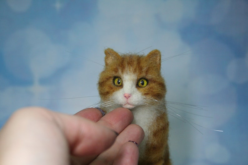 May include: A needle felted cat figurine featuring orange and white fur, pointed ears, and bright yellow eyes. The cat has a small pink nose and long, thin, clear whiskers extending from its muzzle, crafted with a textured, soft appearance.