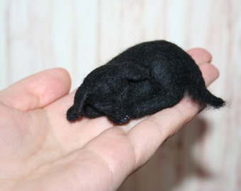 May include: A small, handmade black felted animal figurine, possibly a dog or cat, is resting in a person's open palm. The figurine is made of soft, dark material and has a simple, rounded shape with small ears and a tail.