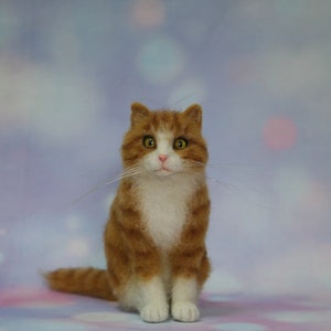 May include: A needle felted sculpture of a ginger and white tabby cat. The cat has orange striped fur on its head and back, a white chest and paws, a small pink nose, and large amber eyes. Long, thin white whiskers extend from its muzzle.