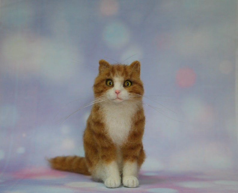 May include: A needle felted sculpture of a ginger and white tabby cat. The cat has orange striped fur on its head and back, a white chest and paws, a small pink nose, and large amber eyes. Long, thin white whiskers extend from its muzzle.