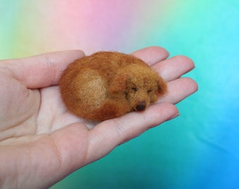 May include: A miniature, handcrafted felted dog figurine in a curled sleeping position. The dog is a warm, reddish-brown color, with closed eyes and a small, dark nose. The figurine is held in an open hand against a soft, colorful background.