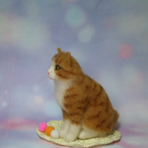 May include: A needle-felted orange and white tabby cat figurine sitting on a small, round, cream-colored crocheted mat. The cat has yellow eyes, a pink nose, and long whiskers. Three small, round, colorful pom-poms in pink, orange, and white sit nearby.