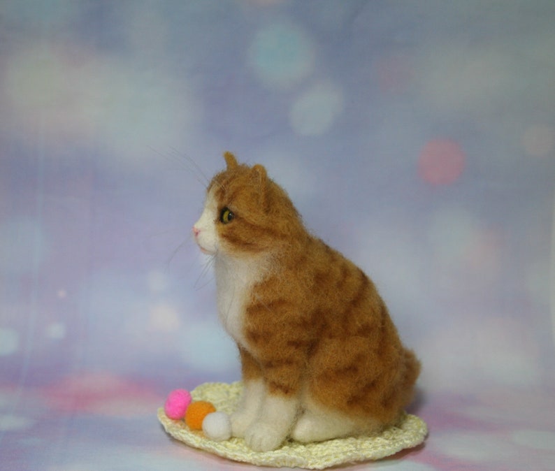 May include: A needle-felted orange and white tabby cat figurine sitting on a small, round, cream-colored crocheted mat. The cat has yellow eyes, a pink nose, and long whiskers. Three small, round, colorful pom-poms in pink, orange, and white sit nearby.