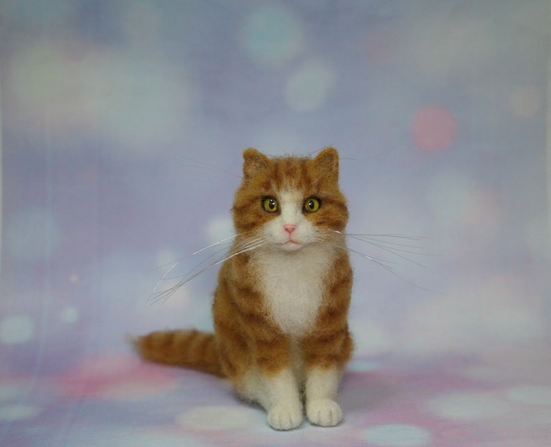 May include: A needle-felted orange and white tabby cat figurine with yellow eyes, a pink nose, and long, thin, clear whiskers. The cat is in a seated position, featuring textured orange fur stripes on its head and back, with a solid white chest and paws.