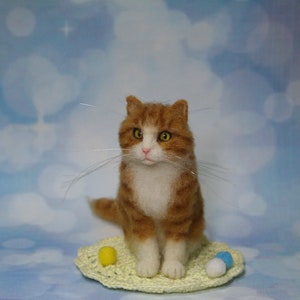 May include: A needle-felted orange and white tabby cat figurine with yellow eyes and long whiskers, sitting on a small, round, pale yellow crocheted mat. The cat is accompanied by three tiny, round, felted balls in yellow, white, and blue.