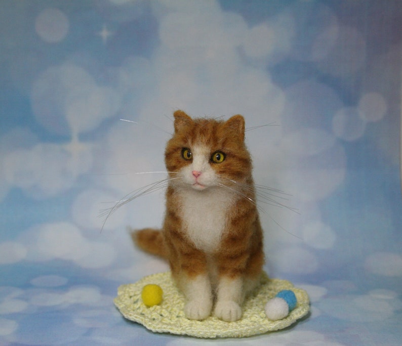 May include: A needle-felted orange and white tabby cat figurine with yellow eyes and long whiskers, sitting on a small, round, pale yellow crocheted mat. The cat is accompanied by three tiny, round, felted balls in yellow, white, and blue.