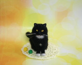 May include: A handcrafted black cat figure with white patches on its chest and paws, and yellow eyes. The cat is positioned on a cream-coloured, crocheted doily with three small, colourful pom-poms.