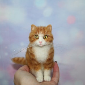 May include: A needle-felted orange and white tabby cat figurine with yellow eyes, a pink nose, and long, thin, clear whiskers. The cat is depicted in a seated position with a textured, wool-like appearance and detailed facial features.