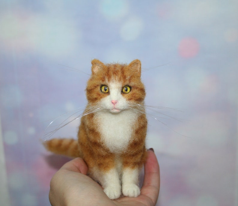 May include: A needle-felted orange and white tabby cat figurine with yellow eyes, a pink nose, and long, thin, clear whiskers. The cat is depicted in a seated position with a textured, wool-like appearance and detailed facial features.