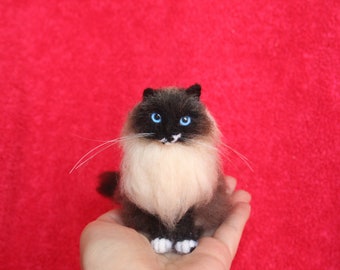 May include: A needle felted cat figurine with white fur, black markings, and blue eyes. The cat is sitting on a hand.