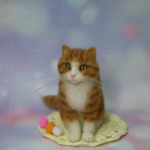 May include: A needle-felted orange and white tabby cat figurine with yellow eyes and long, thin whiskers. The cat sits on a small, round, pale yellow crocheted doily next to three tiny, colorful felted balls in pink, orange, and white.