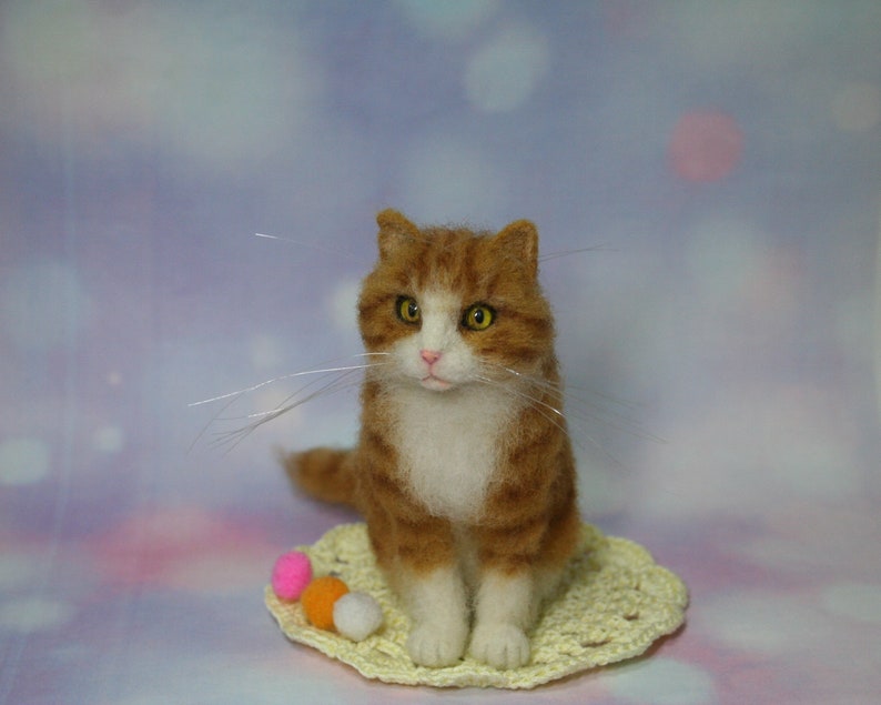 May include: A needle-felted orange and white tabby cat figurine with yellow eyes and long, thin whiskers. The cat sits on a small, round, pale yellow crocheted doily next to three tiny, colorful felted balls in pink, orange, and white.
