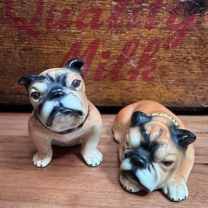 May include: Two ceramic bulldog figurines featuring tan and brown coats with black facial markings. One bulldog is in a standing position, while the other is lying down and wearing a yellow collar with the word "Mom" printed on it.