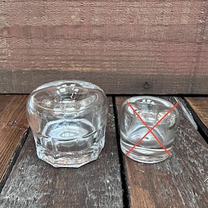 May include: A clear glass candle holder with a thick, faceted base and a smooth, rounded top section featuring a central circular opening for a candle. The glass is transparent and reflects light, showcasing a sturdy, multi-sided bottom design.