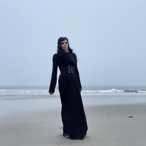 Op de afbeelding: Een vrouw in een zwarte maxi-jurk met een zwart korset en een zwarte choker staat op een zandstrand met de zee op de achtergrond.