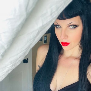 Op de afbeelding: Een vrouw met lang zwart haar en rode lippen kijkt uit achter een wit laken. Ze draagt een zwart, mouwloos topje en een zilveren ketting.