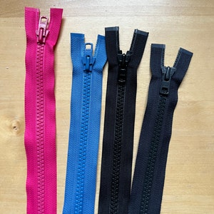 May include: Four zipper closures in different colours: pink, blue, black, and dark grey. The zippers are all open and laid out on a wooden surface.