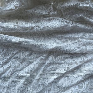 Könnte beinhalten: Nahaufnahme eines weißen Spitzenstoffs mit aufwendigen floralen und geometrischen Mustern. Das zarte Material ist drapiert und erzeugt Schatten und Lichteffekte, die die Textur hervorheben. Die Spitze scheint ein feines Gewebe zu sein, das für Kleider oder andere Kleidungsstücke geeignet ist.