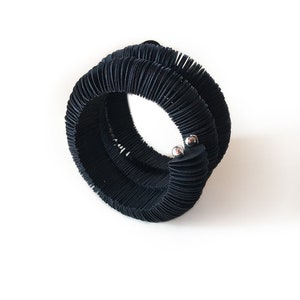May include: A black, coiled bracelet with a textured, ribbed design. The bracelet is made of a flexible material and features two small, silver-colored metal accents. The bracelet is shown against a white background.