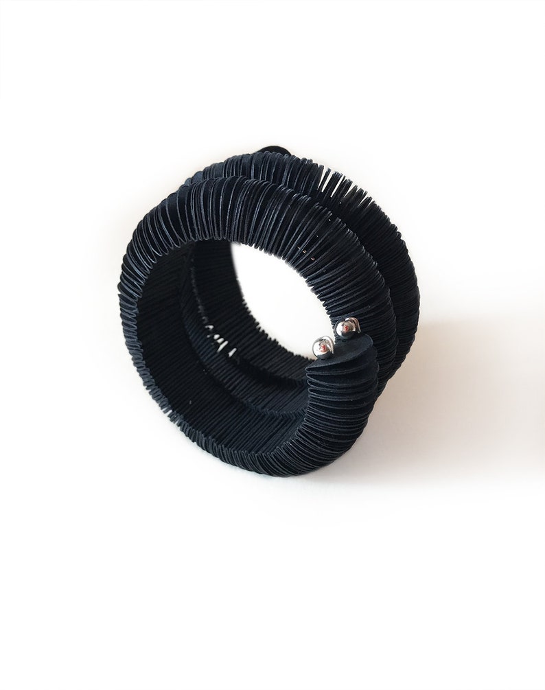 May include: A black, coiled bracelet with a textured, ribbed design. The bracelet is made of a flexible material and features two small, silver-colored metal accents. The bracelet is shown against a white background.