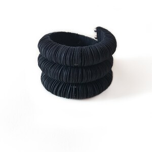 May include: A stack of three black, textured bracelets. The bracelets are made of a series of thin, curved bands, creating a ribbed effect. The top bracelet has a silver clasp. The background is plain white.
