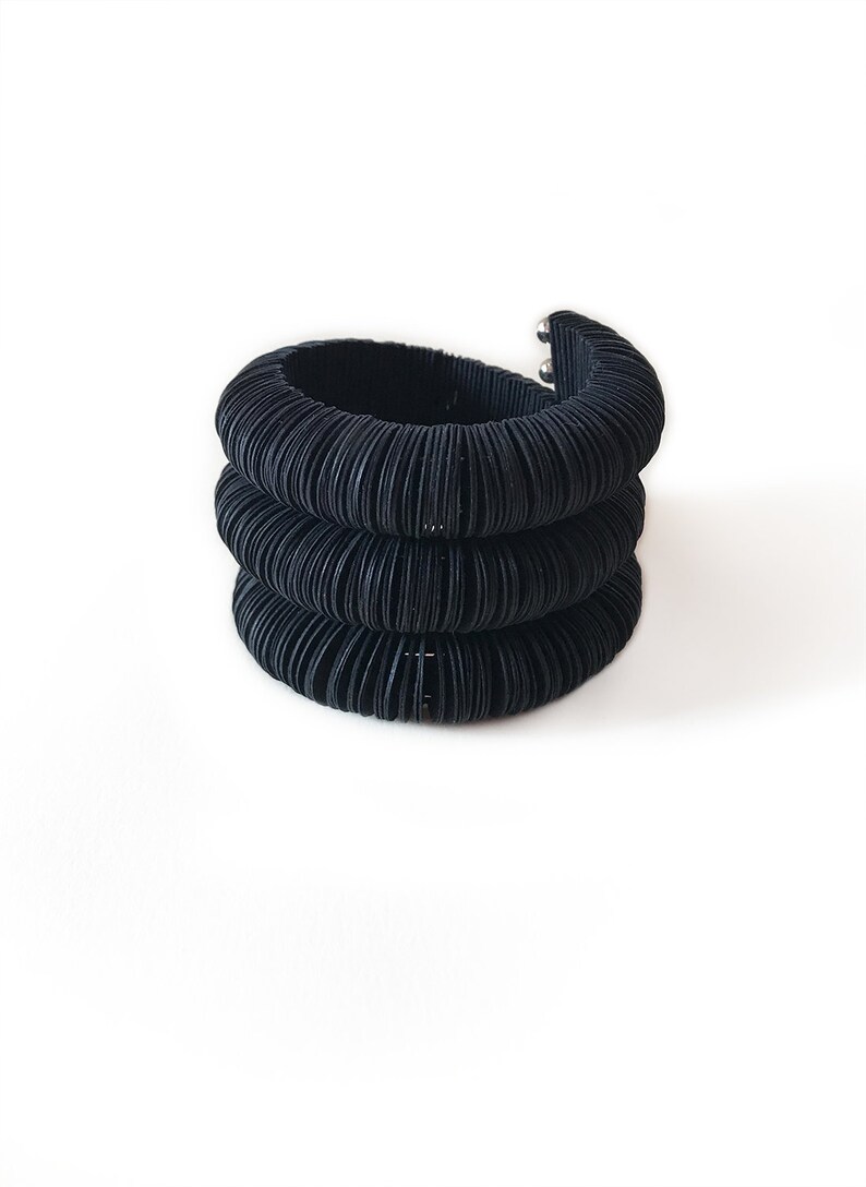 May include: A stack of three black, textured bracelets. The bracelets are made of a series of thin, curved bands, creating a ribbed effect. The top bracelet has a silver clasp. The background is plain white.