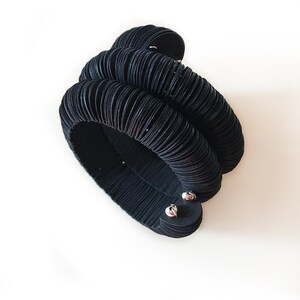 May include: A black, layered bracelet with a unique, textured design. The bracelet appears to be made of multiple stacked, circular elements, creating a bold, sculptural look. The bracelet is secured with two silver-toned clasps.