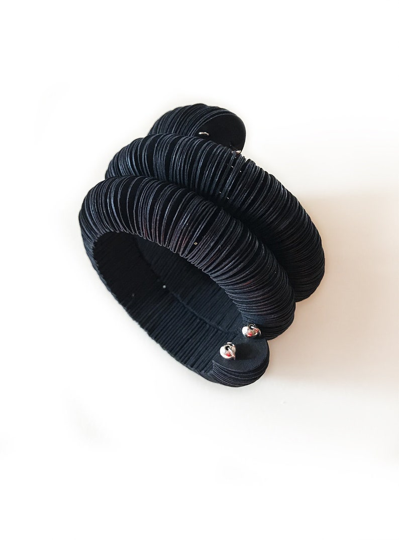 May include: A black, layered bracelet with a unique, textured design. The bracelet appears to be made of multiple stacked, circular elements, creating a bold, sculptural look. The bracelet is secured with two silver-toned clasps.