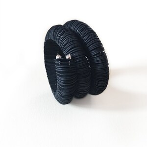 May include: A black bracelet with a unique design. The bracelet is made of multiple stacked, textured bands, creating a layered effect. Two small silver-colored accents are visible on the side. The bracelet is set against a white background.