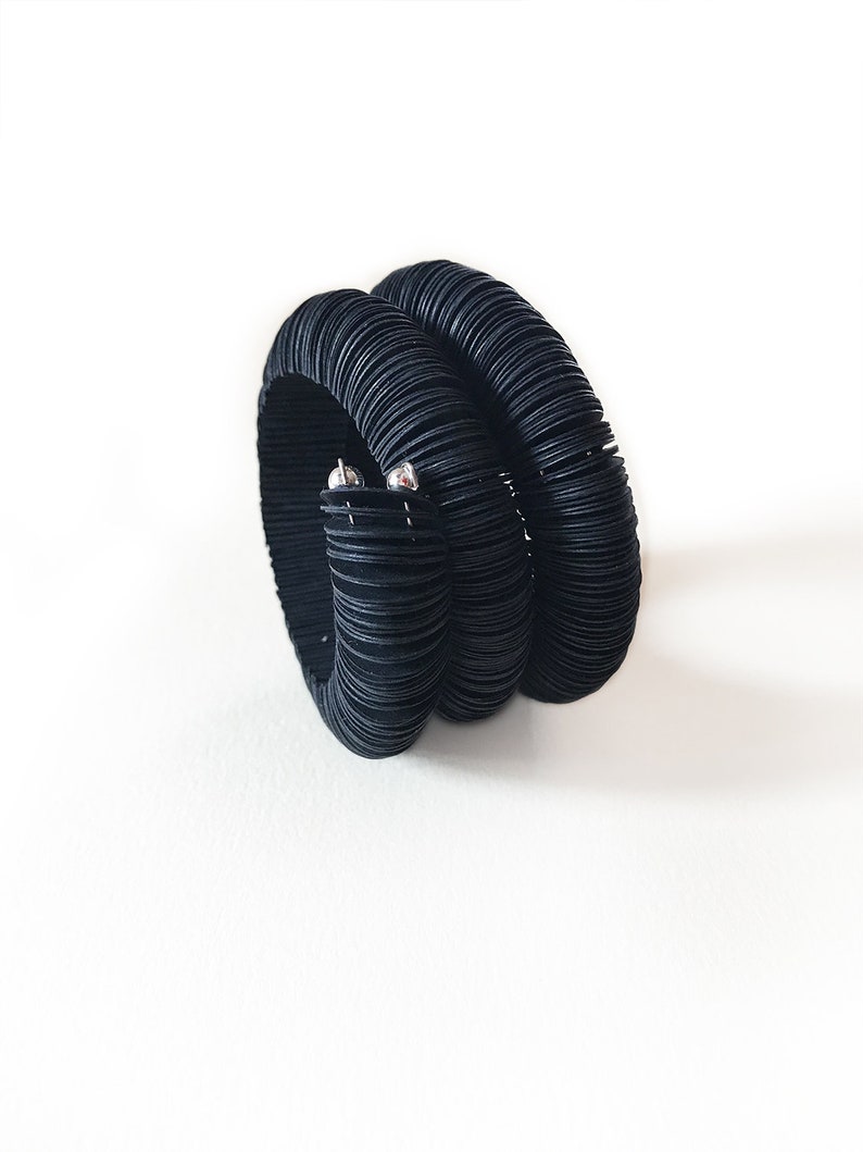 May include: A black bracelet with a unique design. The bracelet is made of multiple stacked, textured bands, creating a layered effect. Two small silver-colored accents are visible on the side. The bracelet is set against a white background.