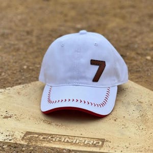 May include: White baseball cap with a brown leather patch featuring the number 7. The brim has a red underside and red stitching. The cap is resting on a baseball base.