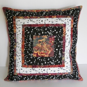 May include: Square decorative pillow with a musical theme. The pillow features a central square with a floral design, surrounded by concentric squares of black fabric with white musical notes, white fabric, and a burgundy border. The edges are also black with musical notes.