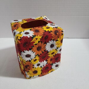 May include: A square tissue box cover featuring a vibrant floral pattern. The design showcases a mix of sunflowers in shades of yellow, orange, red, and white. The cover has a rectangular opening at the top for tissue dispensing.