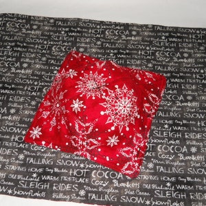 May include: A red fabric hot pad with a white snowflake pattern. The hot pad is on top of a black and white fabric placemat with the words "Sleigh Rides", "Hot Cocoa", "Falling Snow", and "Cozy Blankets" printed on it.
