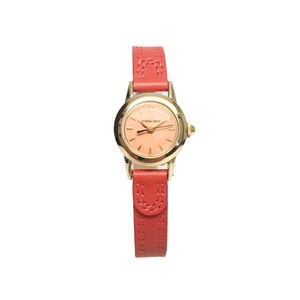 Puede incluir: Un reloj de pulsera dorado con una esfera rosa y una correa de cuero roja. La esfera del reloj tiene el texto "ETERNA MATIC" impreso en ella.