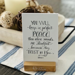 May include: A white card with black calligraphy text that reads "YOU WILL keep in perfect Peace those whose minds are Steadfast because they TRUST in You Isaiah 26:3". The card is standing on a small wooden block with the name "Kristin" written on it.