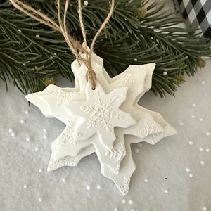May include: White snowflake ornaments with intricate snowflake patterns, strung with twine. The layered design creates a 3D effect. Festive decor for Christmas or winter holidays. The ornaments are displayed with evergreen branches and a gift box.