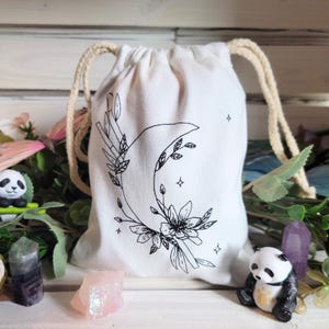 May include: A white cotton drawstring bag with a black and white illustration of a crescent moon, flowers, and stars. The bag is perfect for storing small items like crystals, jewelry, or other trinkets.